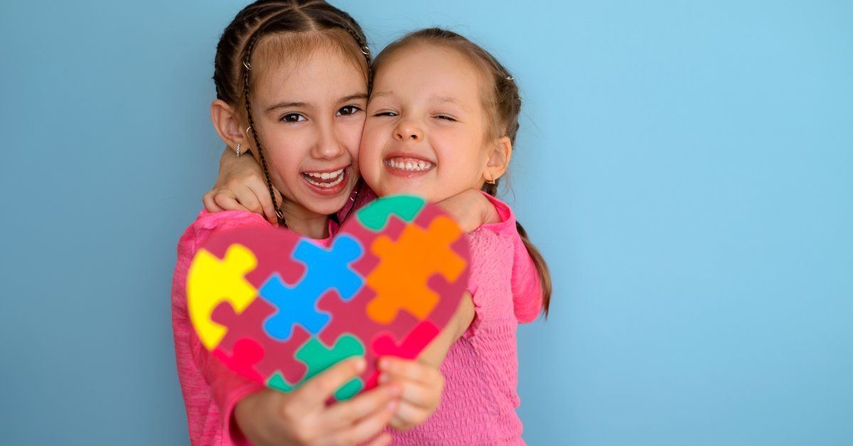 Two young girls smiling and hug while holding out a heart-shaped puzzle containing multi-colored pieces.