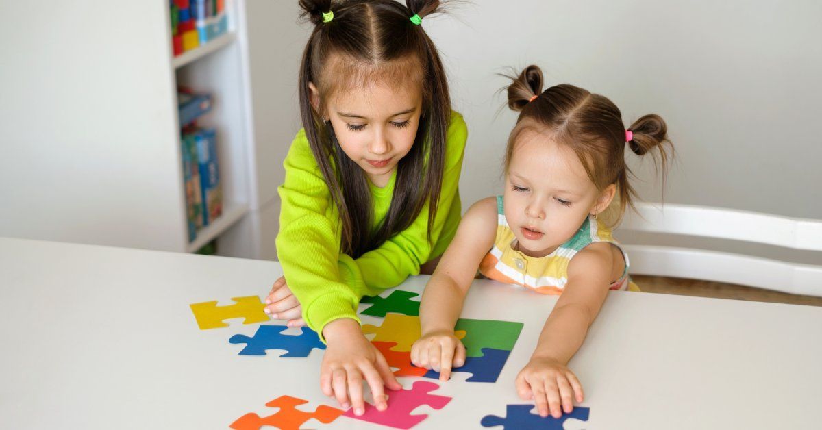 Two young girls standing at a table while arranging colorful puzzle pieces.