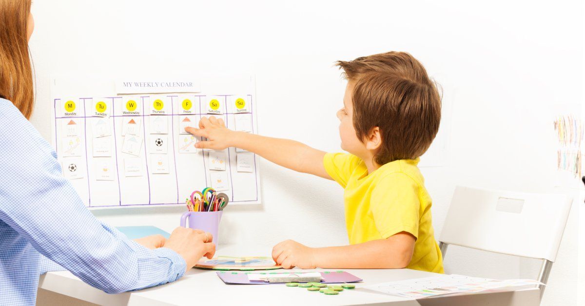 A little boy wearing a yellow shirt sits at a desk while pointing to a calendar that hangs on the wall.