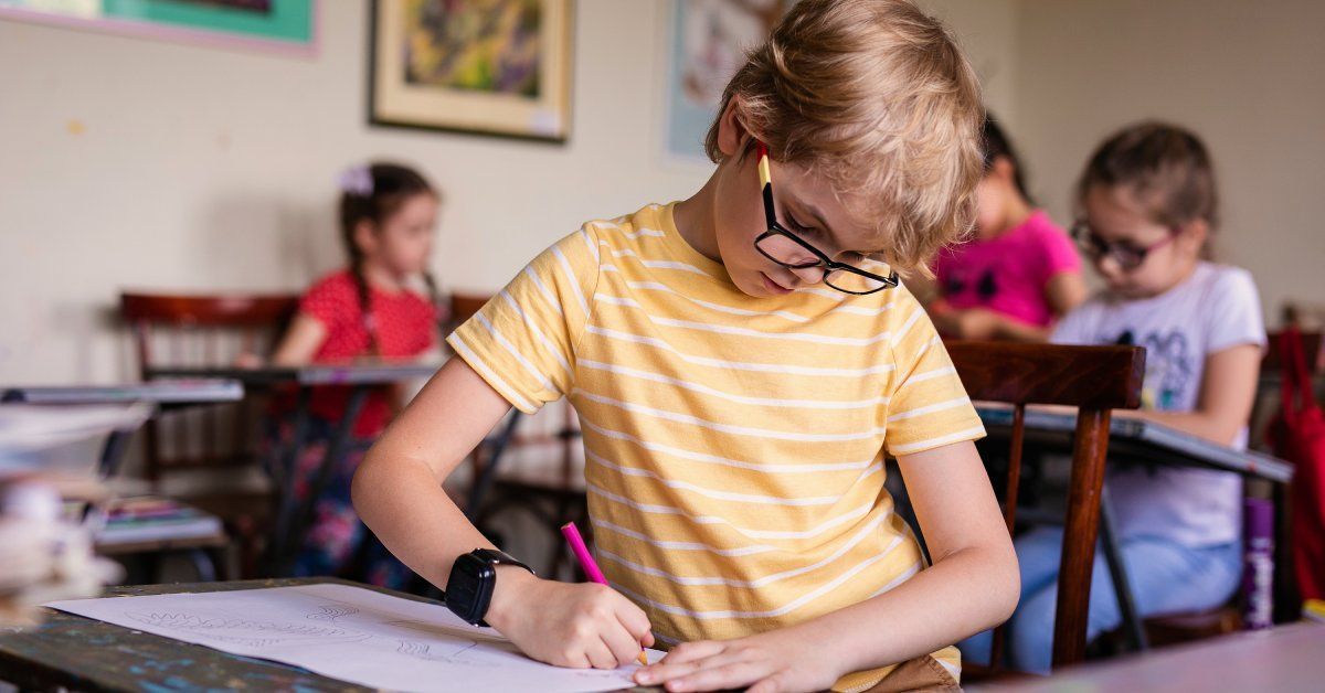 A boy wearing a striped shirt sits at a desk while using a pen to color. Other students are coloring