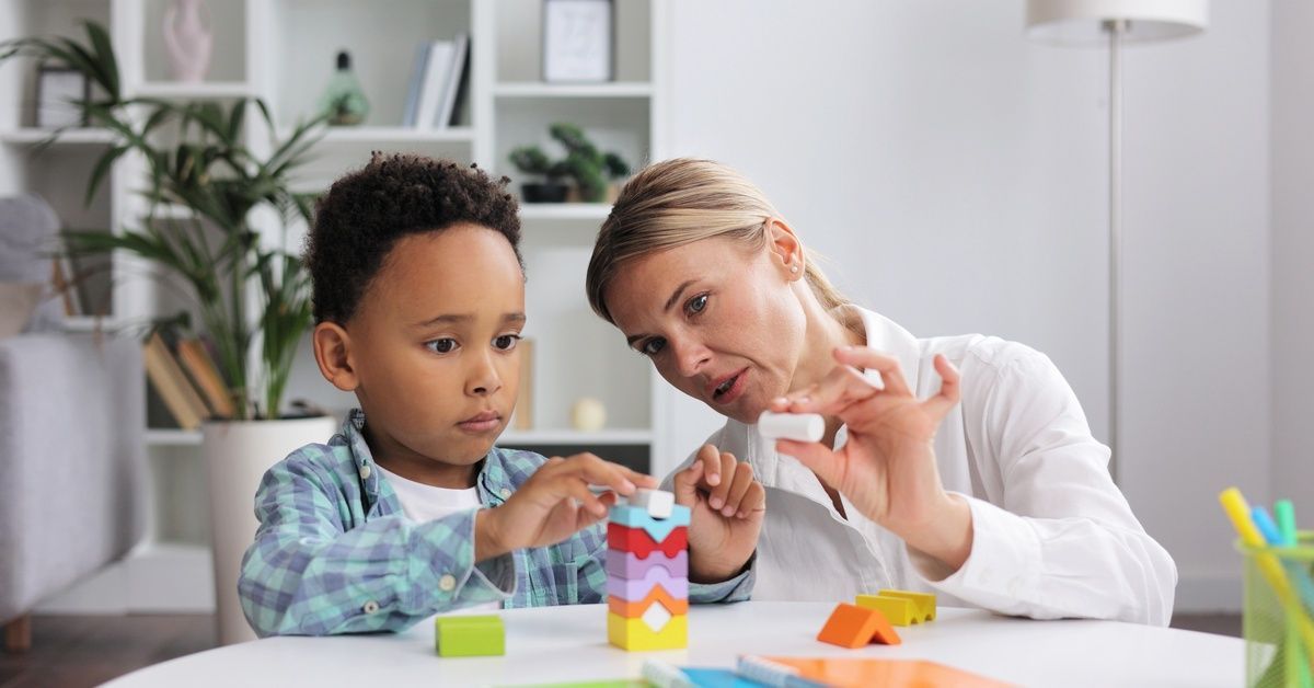 A little boy sitting at a table while stacking multi-colored blocks on top of one another while a woman sits nearby.