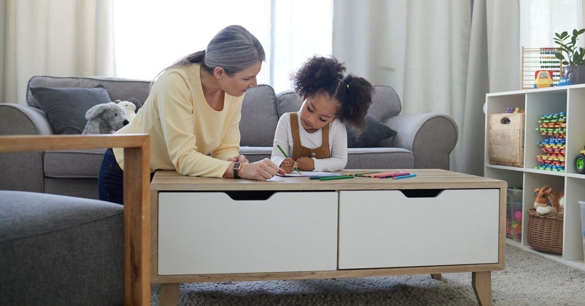 A little girl sitting on a couch near an older woman, while using colored pencils to draw and color on a piece of paper.