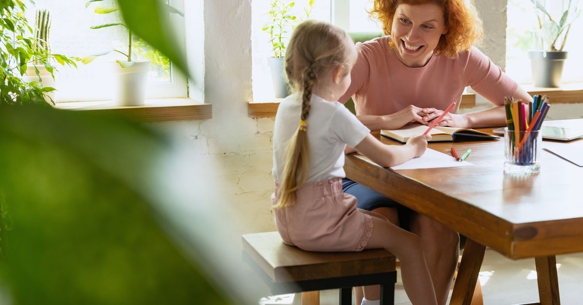 A woman at a small table next to a girl who's drawing with a colored pencil.