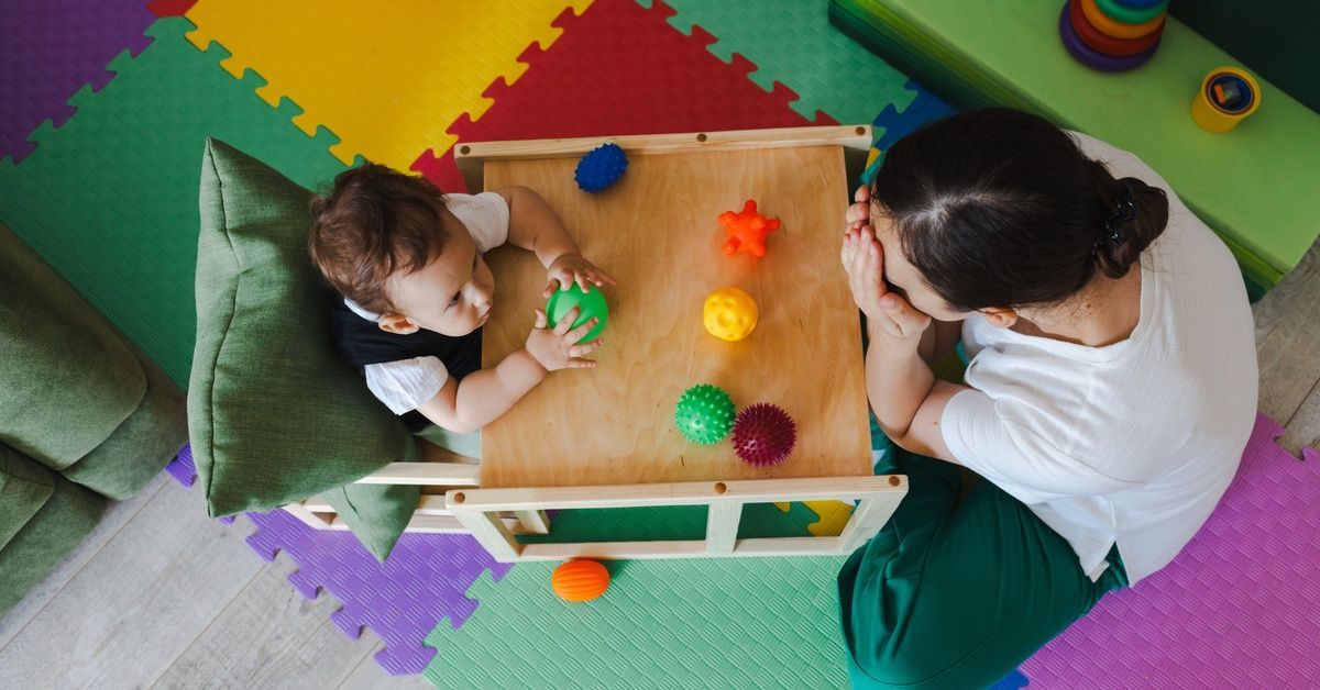 A top-down shot of a baby in a wooden high chair with toys on the table. A woman plays peekaboo across from him.