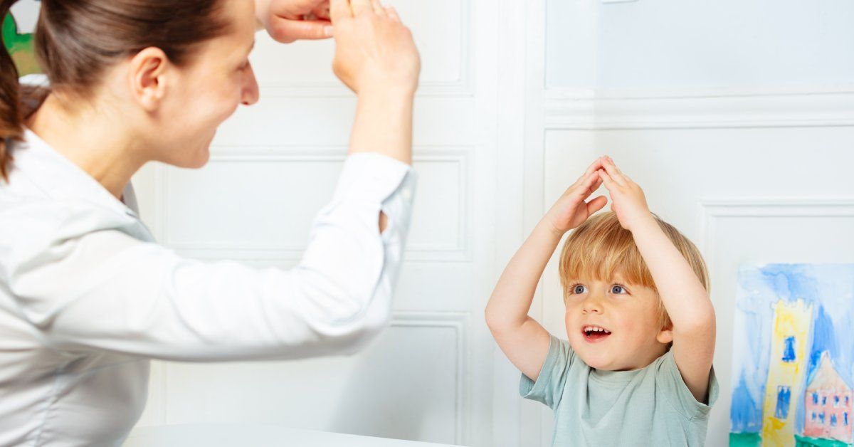 A little boy sits across from his therapist and mimics her holding her arms up.
