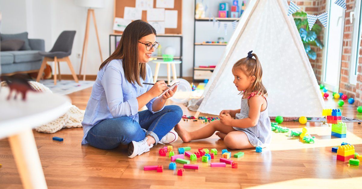 A therapist and child sit in a room full of toys as the therapist writes down her observations.