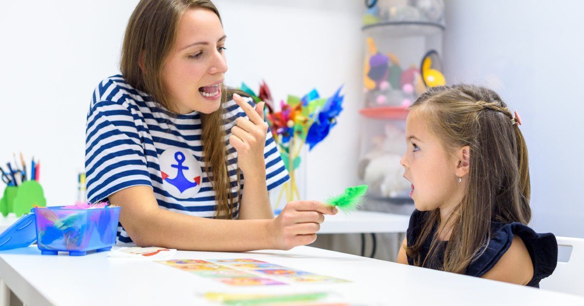 A mother and child sit at a desk as the mother demonstrates proper tongue placement when pronouncing sounds.