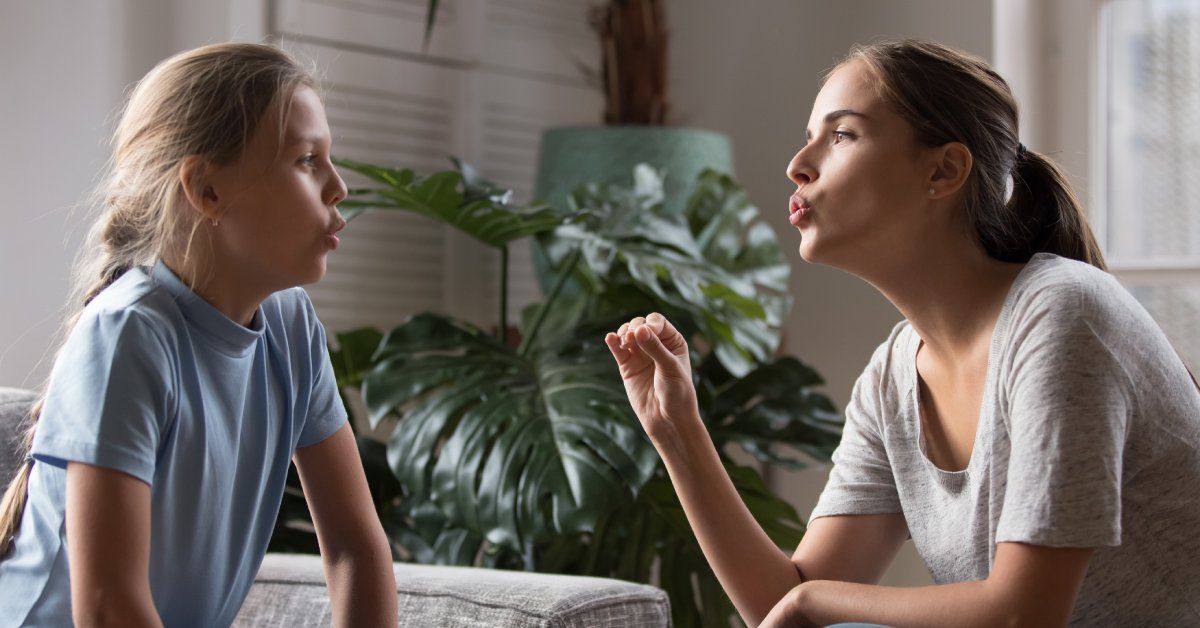 A child and mother sit in the living room as the mother demonstrates proper lip formations for pronouncing different sounds.