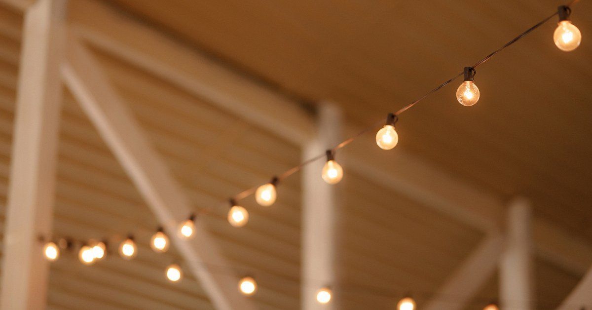 A dimly lit room has warm colored string lights hanging from the ceiling. There are windows with shutters in the background.