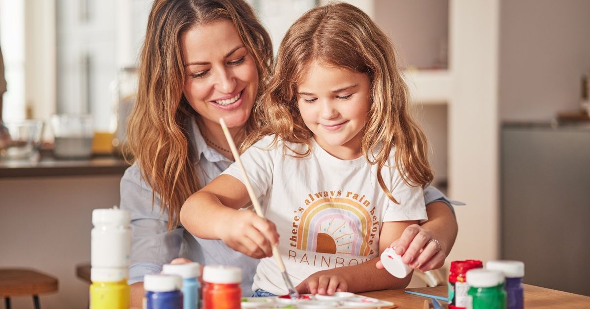 A mother and her child sit at the kitchen table as the mother helps the child dip a paintbrush into paint.