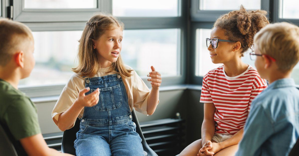 A girl sits in a chair near three classmates and tells them a story. She gestures with her hands while talking.