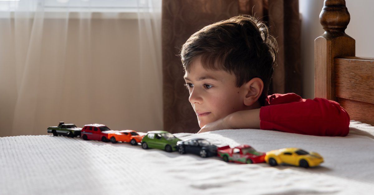 A young boy wearing a red shirt looks intensely at his seven toy cars lined up perfectly on his bed.