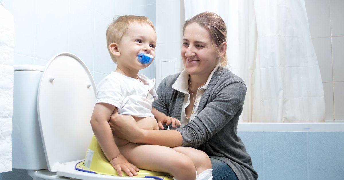 A young child sits on a yellow potty seat resting on a toilet. The child's mother supports and praises the child.