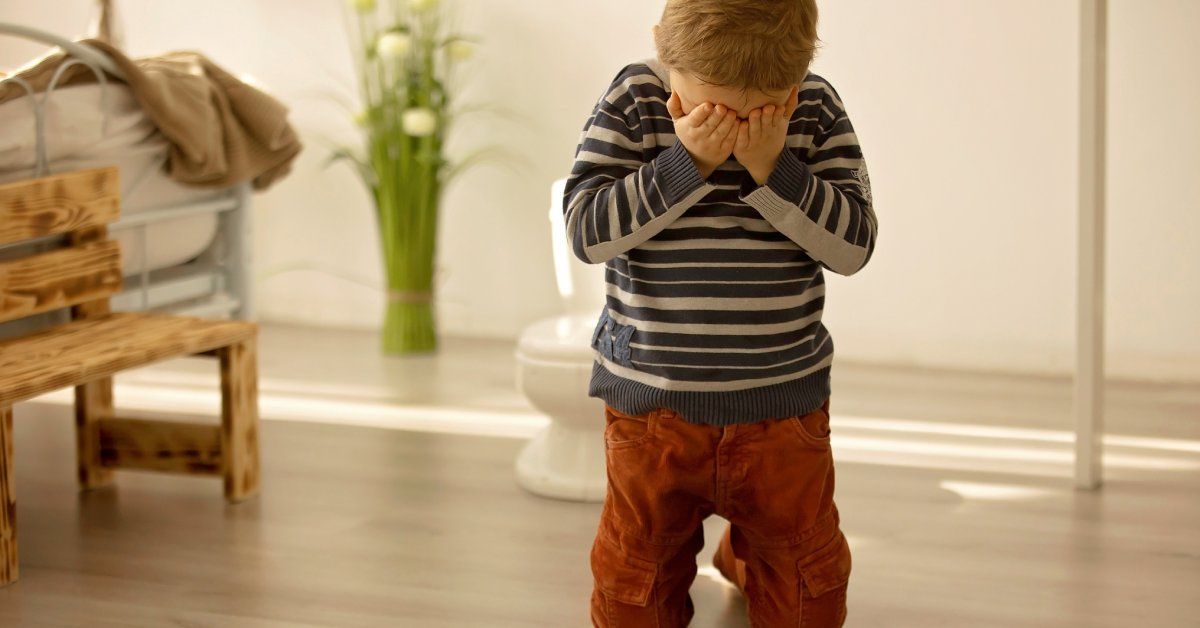 A young boy kneels on the floor, covering his face with his hands.