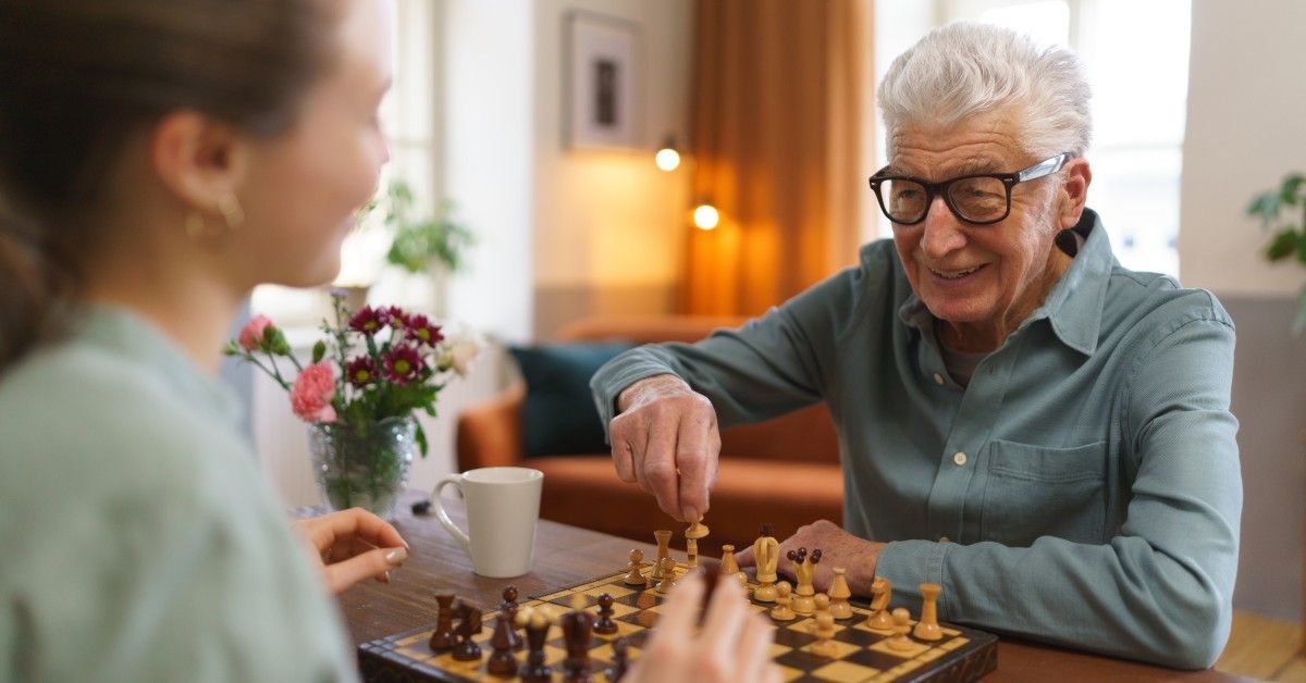 An older man smiles while playing chess with a younger woman at a wooden table. A vase of flowers and a mug sit nearby.