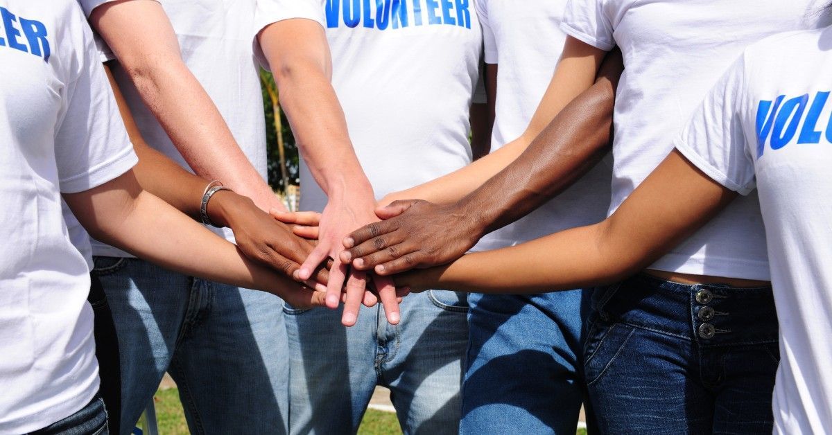A diverse group of volunteers wearing white shirts and jeans stack hands in unity while outside.