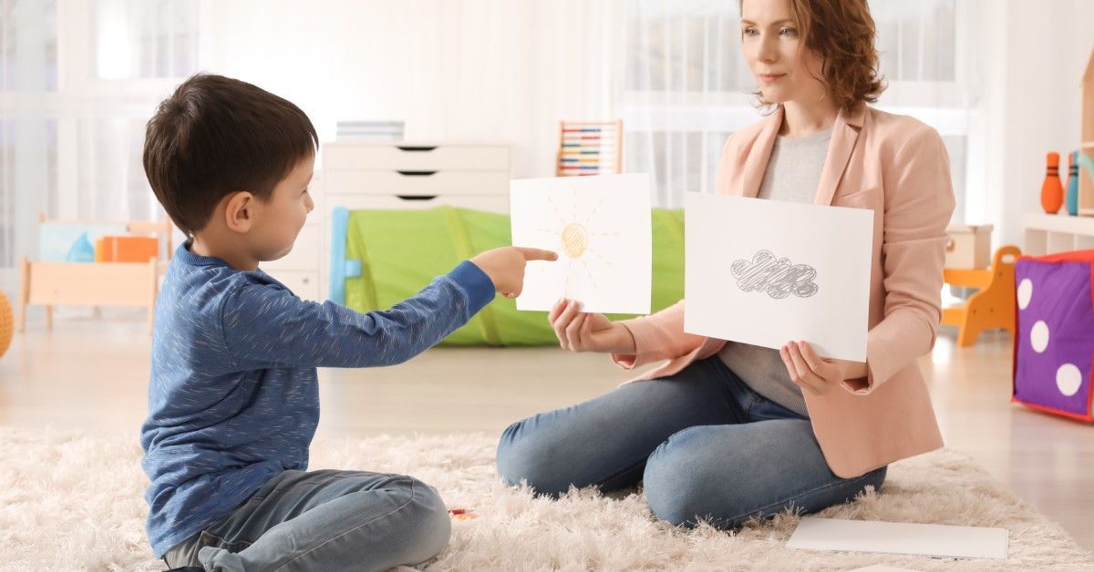 A woman shows sun and cloud cards to a boy who points at the sun card. They sit in a bright room with toys and shelves.