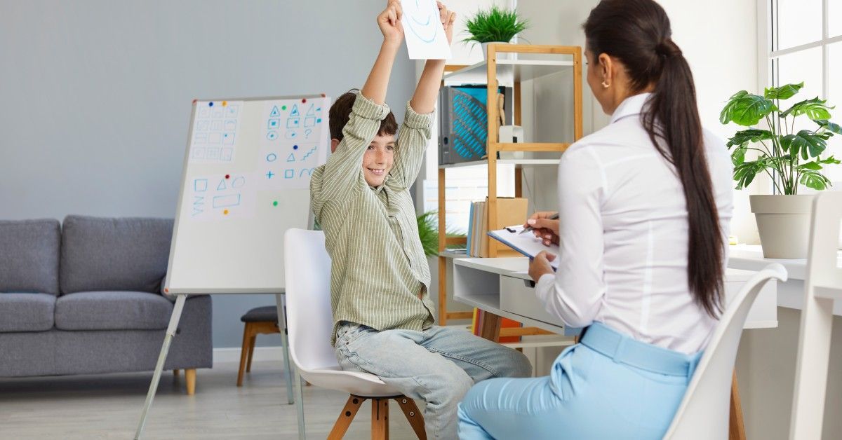 A boy with autism shows a smiling face drawing to an ABA therapist in a bright ABA therapy center.