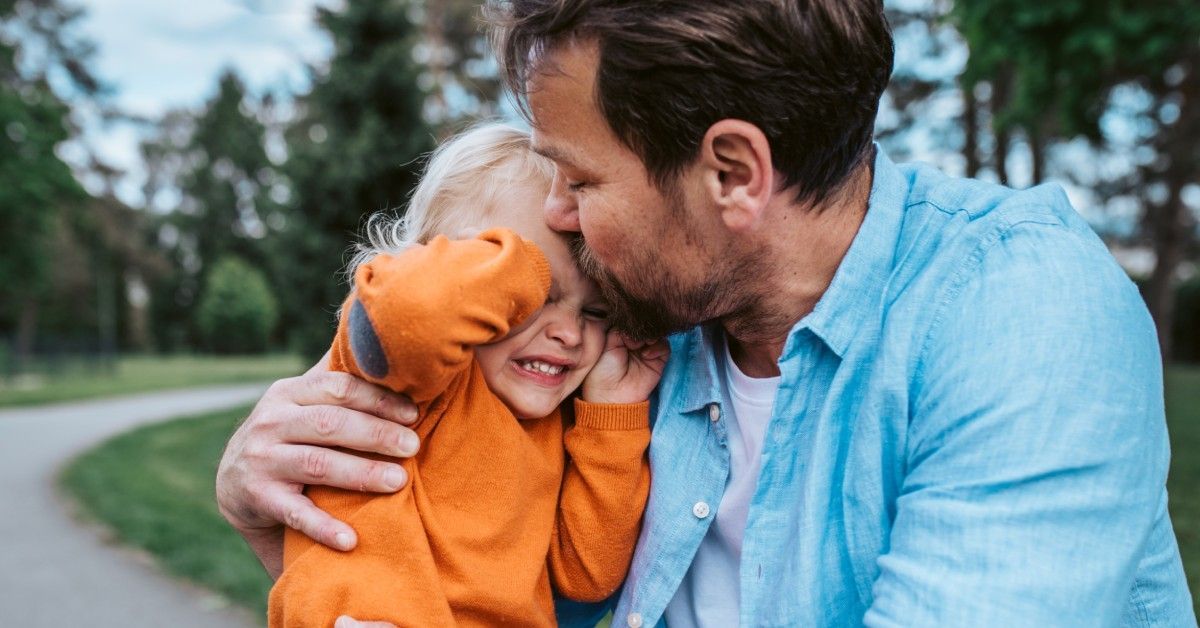 A father embraces and kisses his upset child on the forehead in a park. The child covers their eyes.