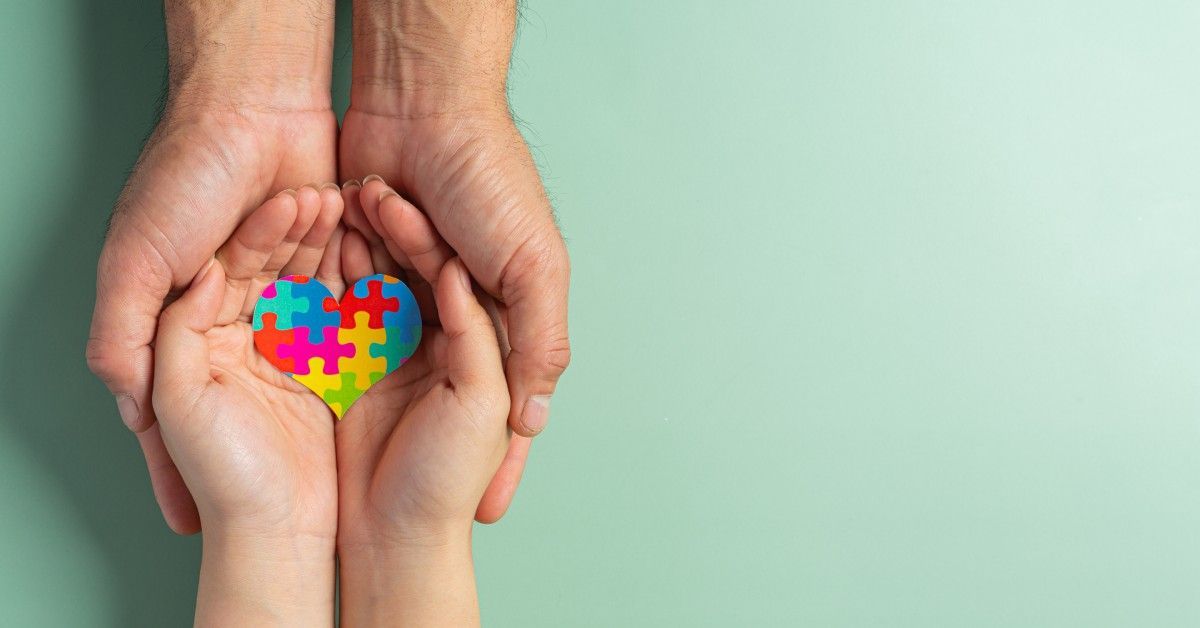 Adult hands gently cradle a child's hands holding a colorful heart-shaped puzzle, symbolizing autism