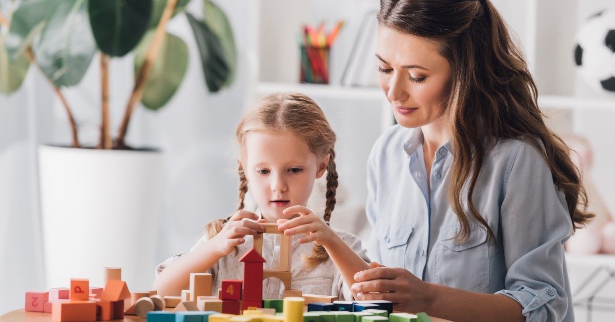 A mother and young girl build with colorful wooden blocks on a round table in a bright, child-friendly room.