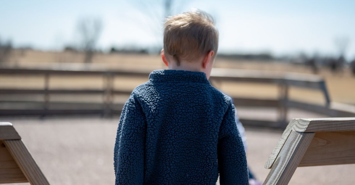 A child in a blue fleece jacket stands outdoors, facing a wooden fence. Trees and a bright sky are blurred in the background.