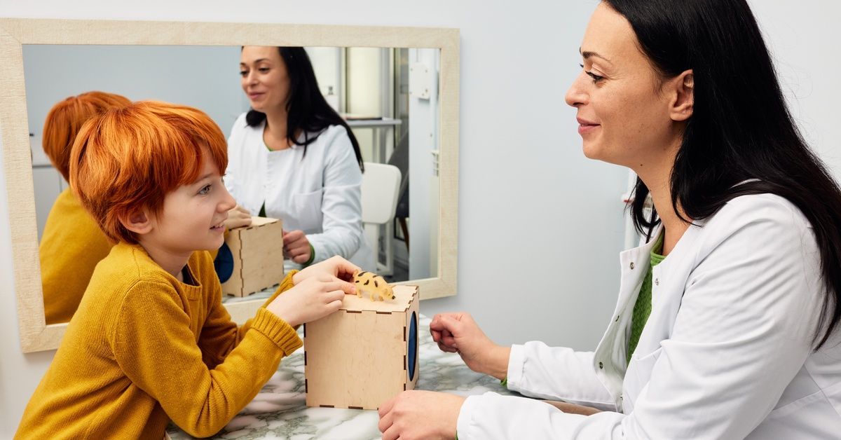 A therapist wearing a white coat and child wearing an orange shirt sit at a table with wooden boxes at a therapy center.