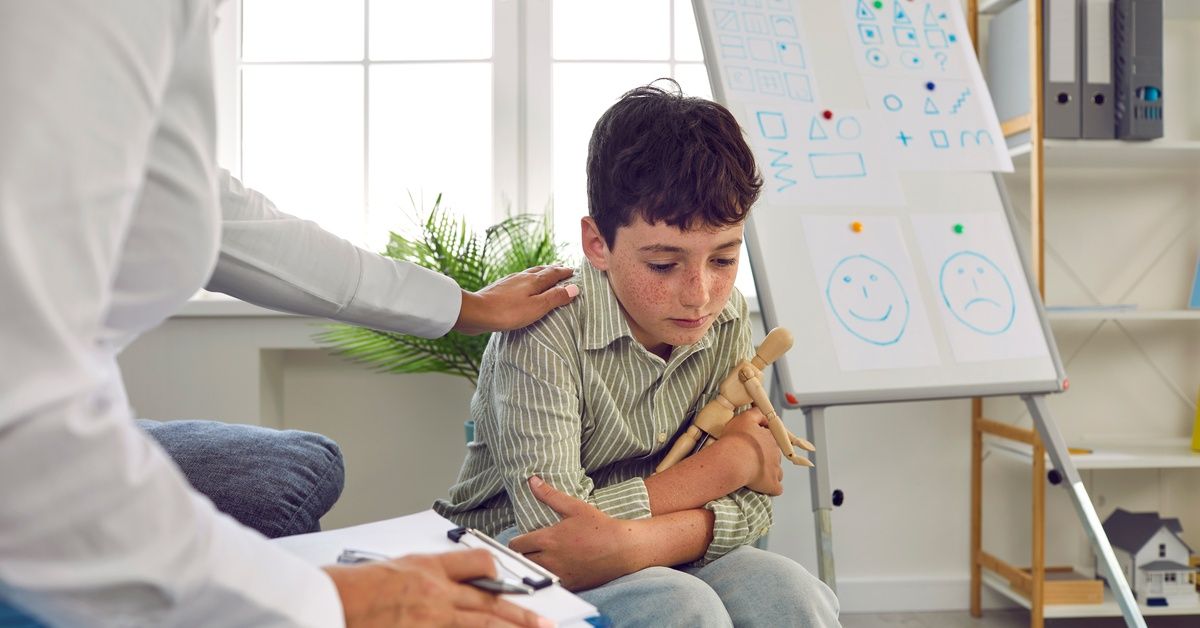A therapist comforts a sad boy holding a wooden figure. A whiteboard with shapes and faces is visibl