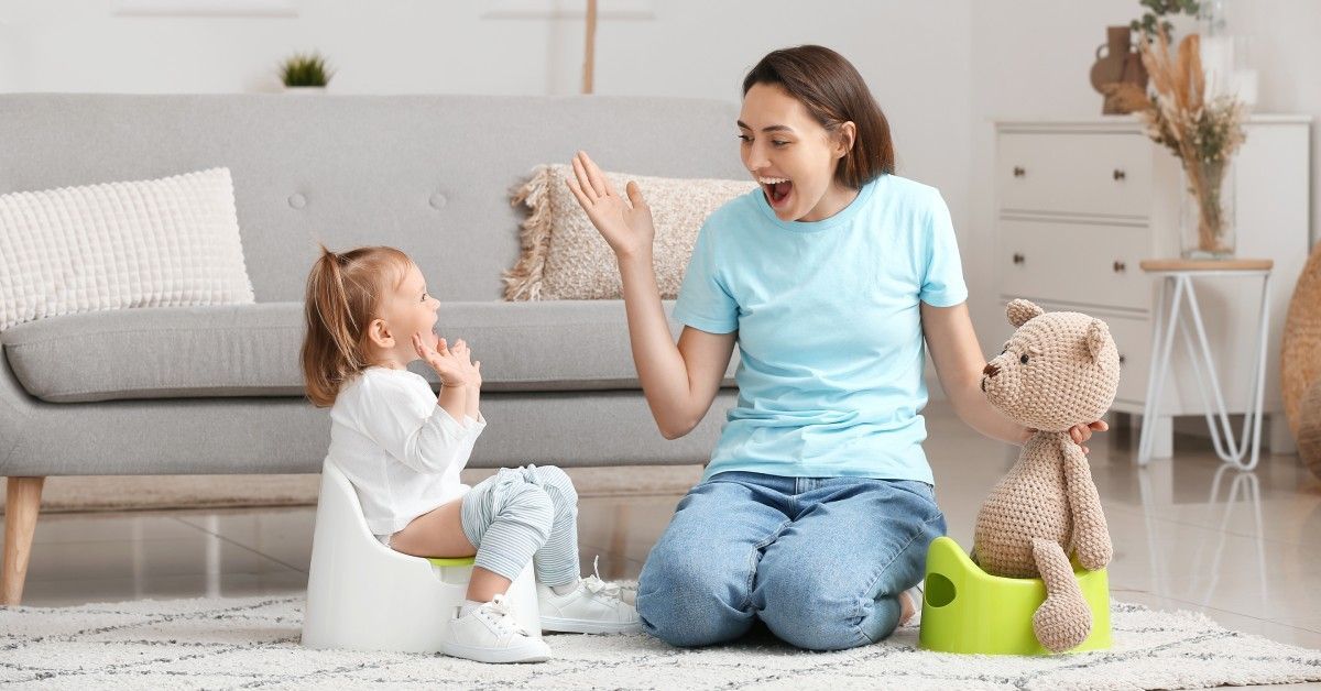 A woman encourages a child sitting on a white potty in a bright living room. A teddy bear sits on a second potty nearby.