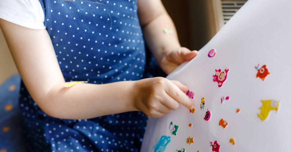 A child wearing a blue polka-dot dress plays with colorful stickers on a large white sheet of paper.
