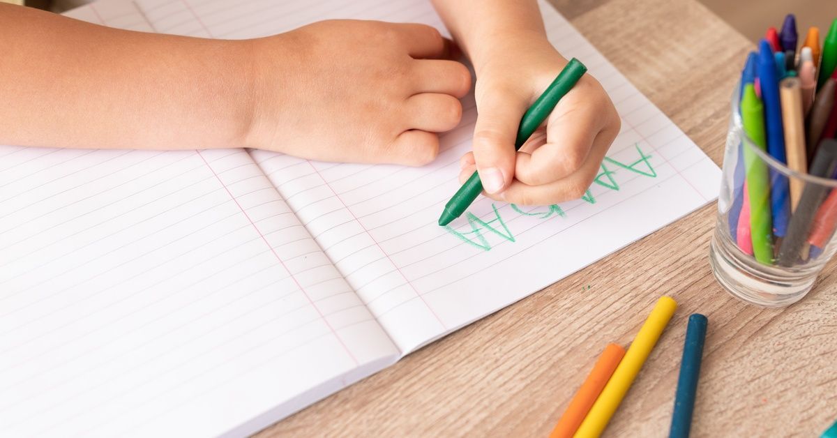 A child writes letters with a green crayon in a lined notebook on a wooden table, surrounded by colorful crayons.