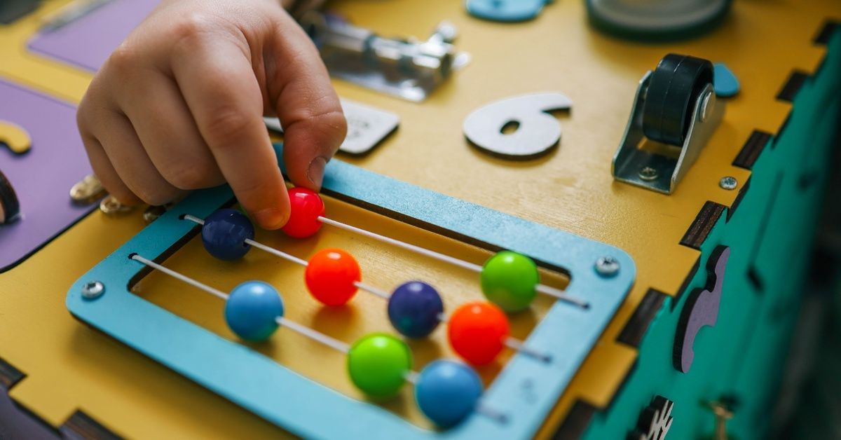 A child's hand pushes a red bead on a colorful abacus attached to a yellow busy board.