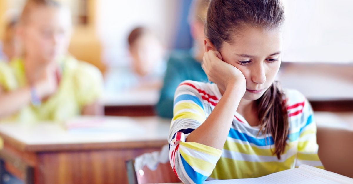 A young girl in a striped shirt focuses on her notebook in a softly lit classroom, with blurred students in the background.