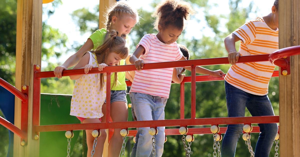Four children walk on a red chain bridge at a playground. Burred green foliage is in the bright background.