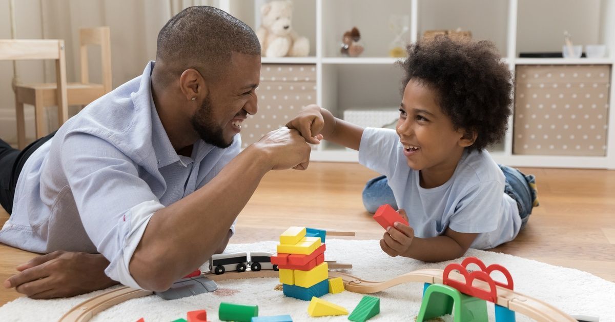A smiling parent and and child lying on a rug fist-bump each other. Colorful toys and a train lie on the ground nearby.