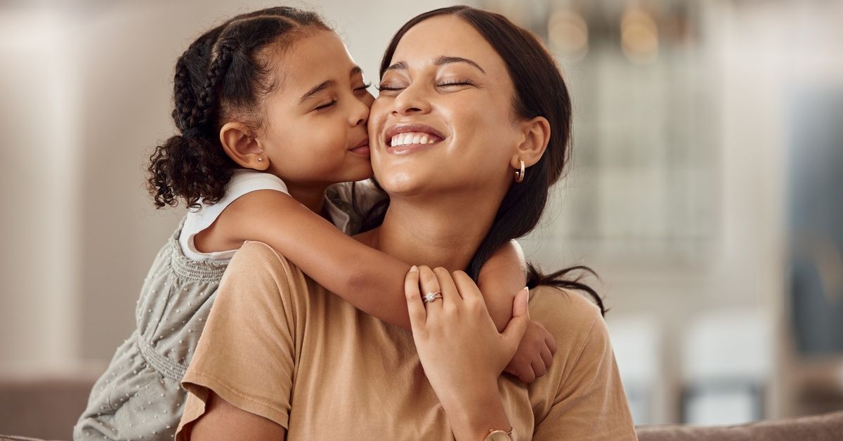 A daughter hugs her mother from behind. The daughter kisses her smiling mother’s cheek.