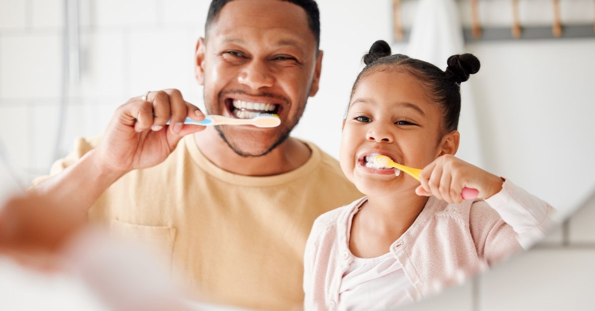 A father and his young daughter smile while brushing their teeth together in a brightly lit bathroom.