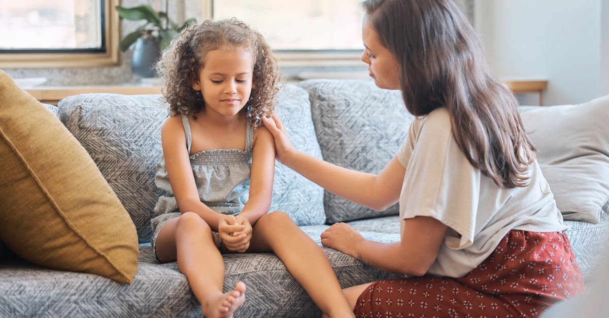 A young girl sits on a patterned sofa, looking down with clasped hands, as her mother gently comforts her.