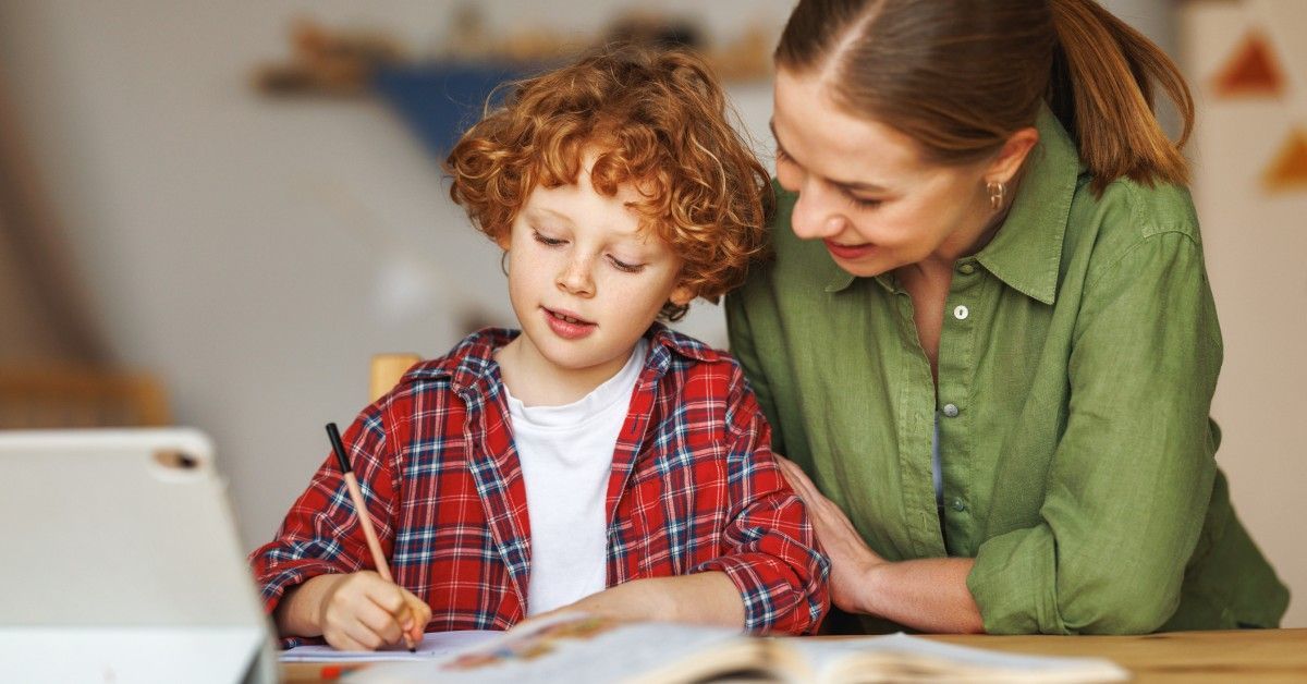 A young boy writes at a table with a book and tablet, while his mom leans in supportively, guiding his work.