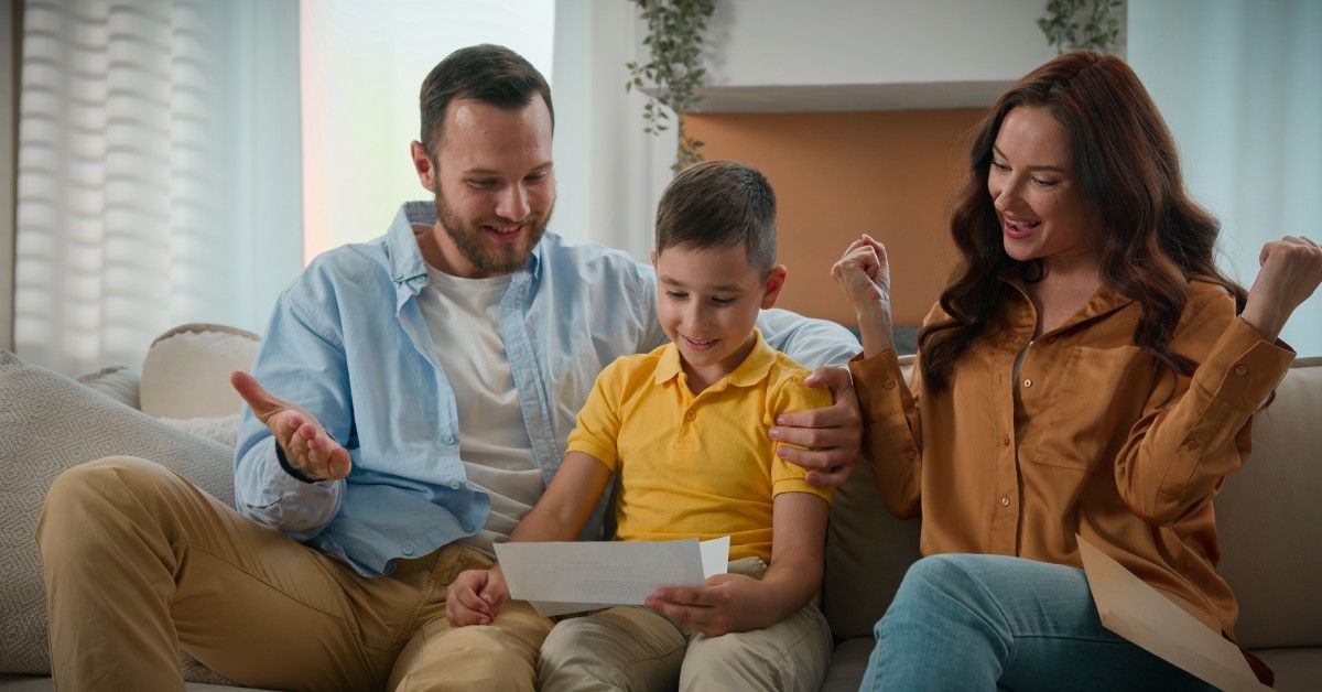 A family sits on a sofa as a young boy holds his test results from school. Both parents smile and celebrate the moment.