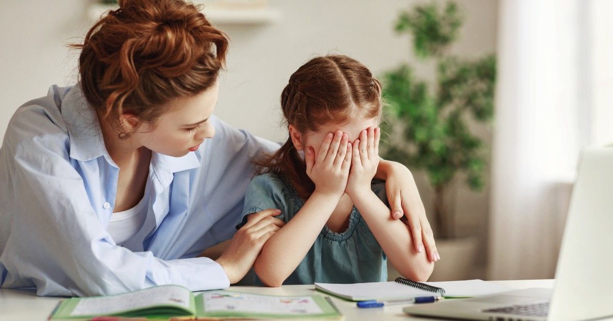A mother comforts her daughter at a table with books, pencils, and a laptop during a study session f