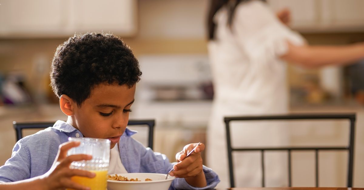 A young boy wearing a blue shirt sits at the kitchen table to eat a bowl of cereal and drink orange juice.