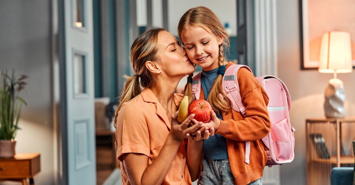 A mom crouches next to her young daughter and gives her a kiss on the cheek before she leaves for sc