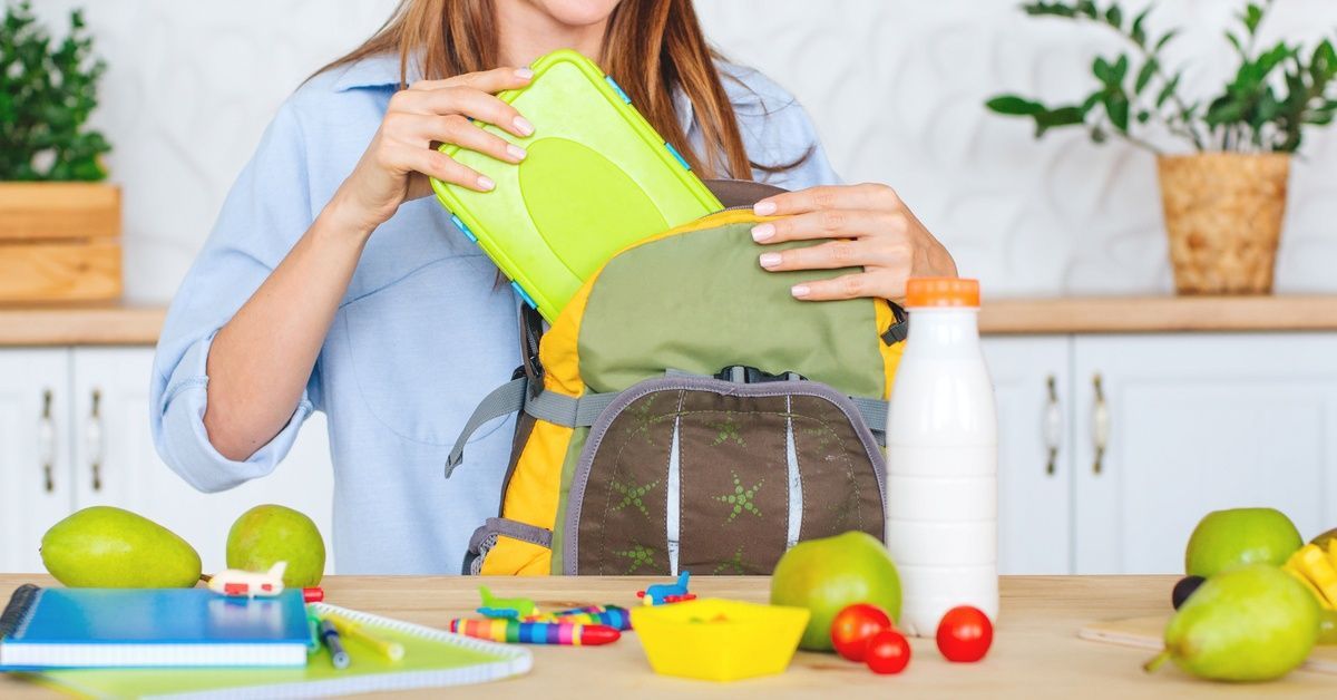 A mom stands at the kitchen counter as she packs her child's school stuff into their backpack before they leave for school.