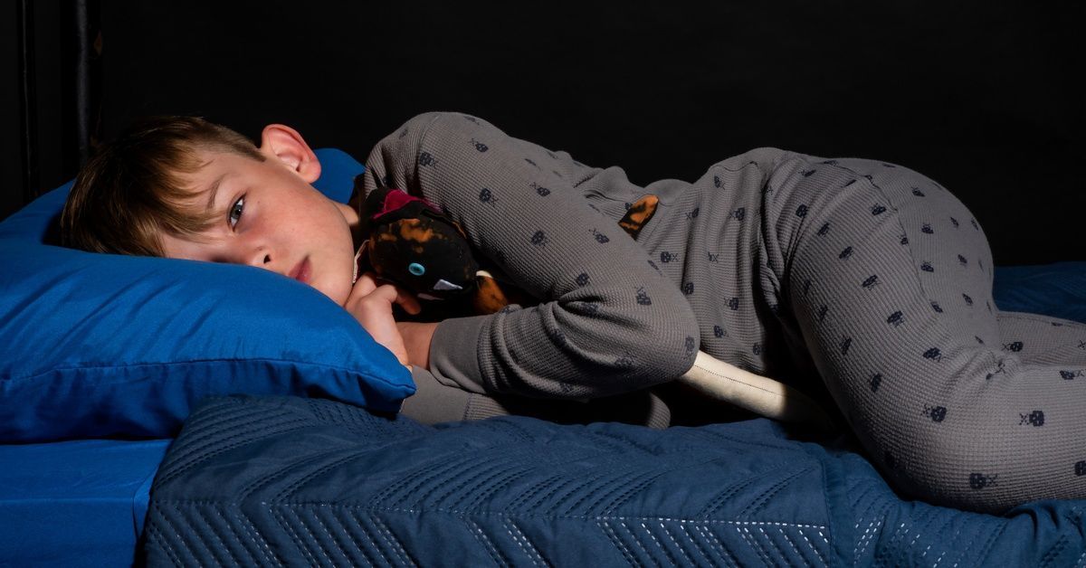 A young boy wearing gray pajamas holds his stuffed animal as he lays awake in bed, struggling to fall asleep.