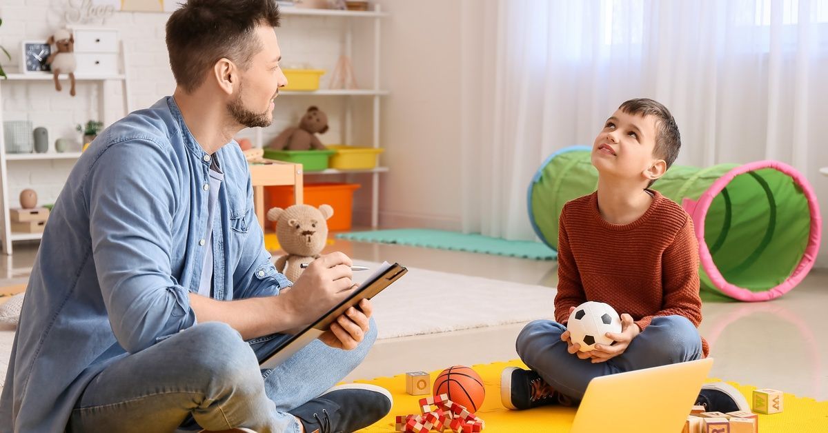 An ABA therapist holding a clipboard sits in a colorful therapy room next to a young boy with autism.