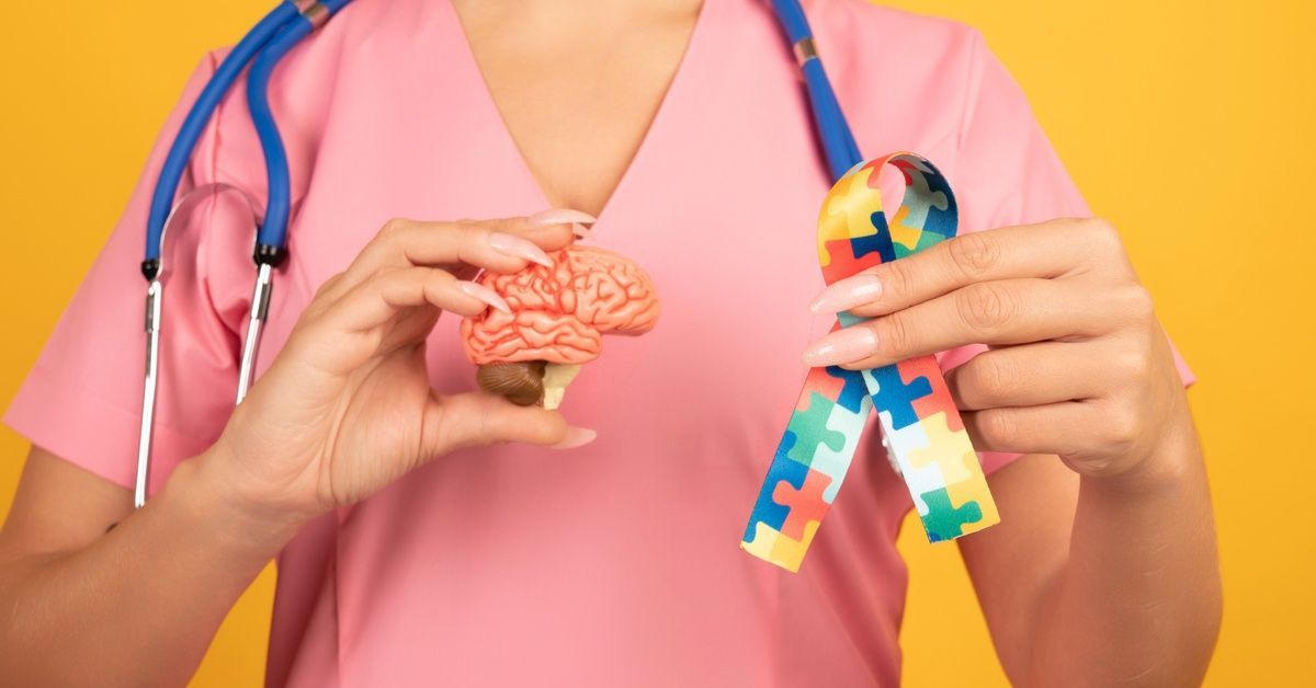 A medical professional wearing pink scrubs holds a model brain and colorful autism awareness ribbon.
