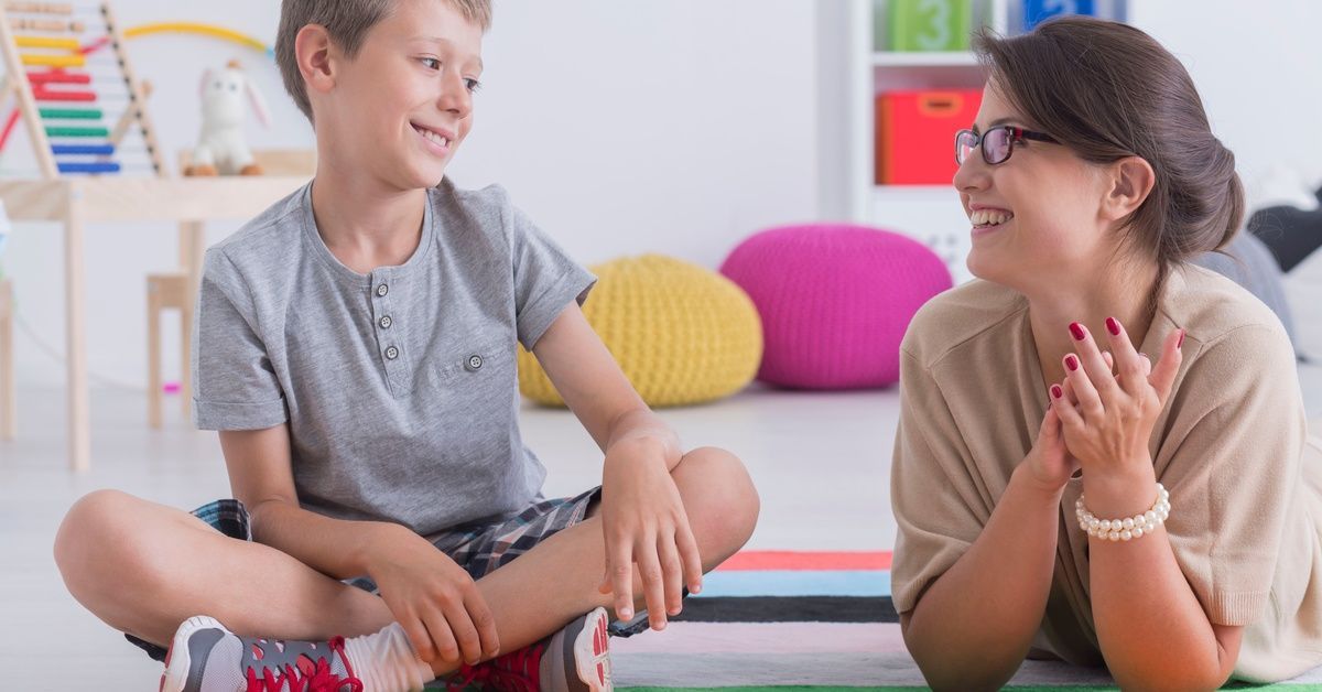 A smiling boy sits on a playroom rug as a therapist lies beside him, clapping and making eye contact.