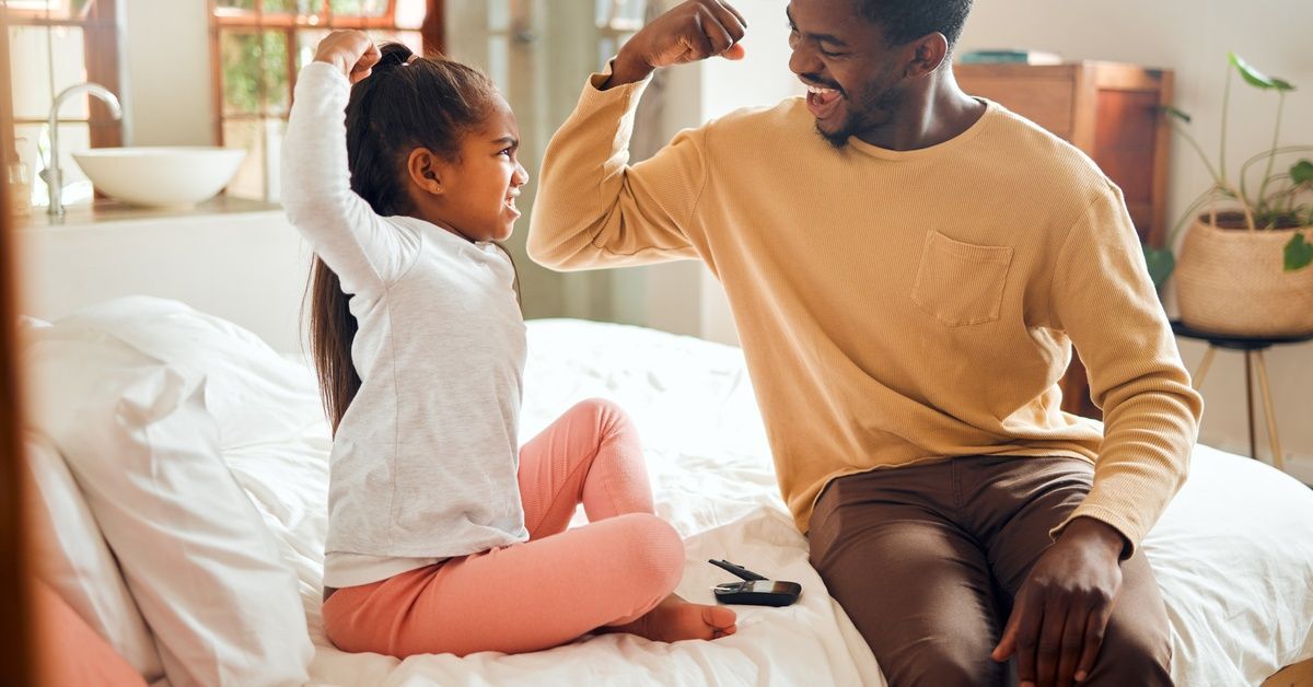 A father and young daughter sit on a bed and smile as they flex their arms together in a playful moment.