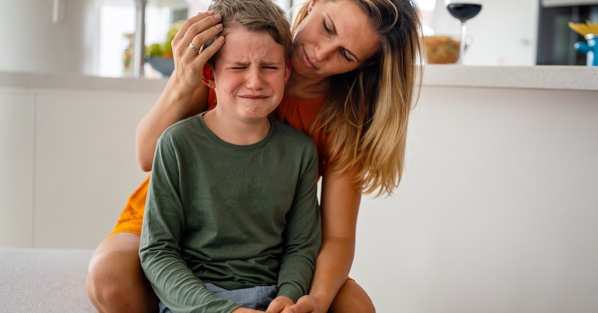 A mother comforts her crying son in their kitchen. She leans over, gently touching his head.